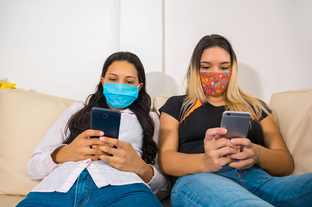 Amigos entediados usando máscaras faciais e telefones inteligentes sentados em um sofá na sala de estar
 - Foto, Imagem