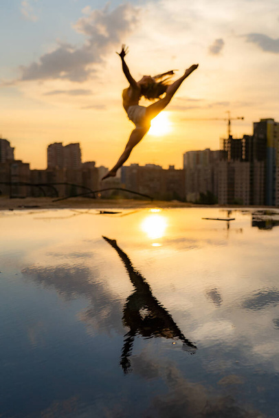 Female flexible dancer jumping during sunset on cityscape background with reflection in the water. Concept of freedom and happiness  - Fotografie, Obrázek