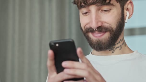 A close-up view of a happy relaxed young hipster man with earbuds is messaging on his smartphone sitting in the living room at home - Materiaali, video