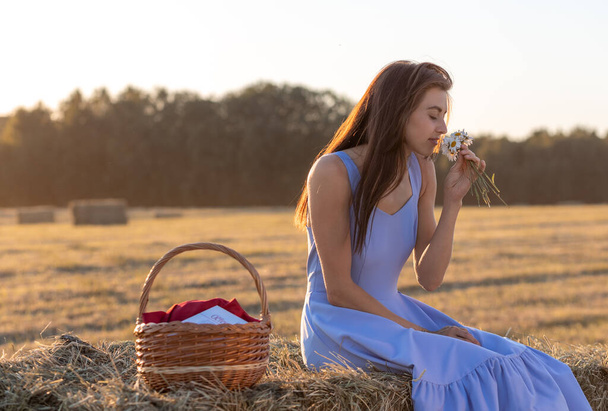 A young woman in a blue dress with a bouquet of daisies is sitting on a haystack in a mown field - Fotografie, Obrázek