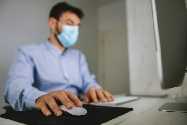 Rückansicht eines Angestellten, der in seinem Büro sitzt, Maske auf dem Gesicht trägt und während des Coronaausbruchs Laptop benutzt. - Foto, Bild