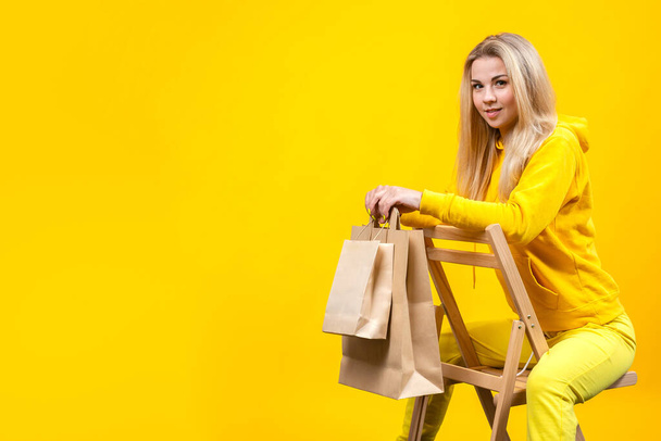 Retrato de jovem mulher loira caucasiana agradável com sacos ecológicos de papel em terno esportivo amarelo, sentado em uma cadeira de madeira, isolado no fundo amarelo, olhando para a câmera. Espaço de cópia
. - Foto, Imagem