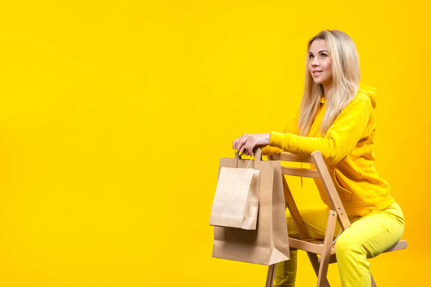 Portrait de jeune jolie femme blonde caucasienne avec des éco-sacs en papier en costume de sport jaune, assise sur une chaise en bois, isolée sur fond jaune. Espace de copie. - Photo, image