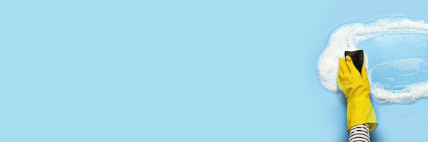 Mano en un guante de goma amarilla sostiene una esponja de limpieza y limpia una espuma jabonosa sobre un fondo azul. concepto de limpieza, servicio de limpieza. Banner. Piso tendido, vista superior. - Foto, imagen