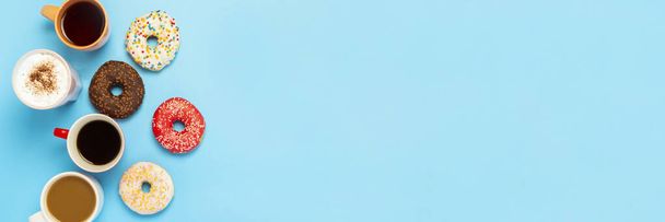 Sabrosas rosquillas y tazas con bebidas calientes, café, capuchino, té sobre un fondo azul. Concepto de dulces, panadería, pastelería, cafetería, reunión, amigos, equipo amable. Banner. Piso tendido, vista superior. - Foto, Imagen