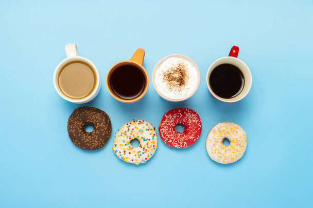 Lekkere donuts en kopjes met warme dranken, koffie, cappuccino, thee op een blauwe achtergrond. Concept van snoep, bakkerij, gebak, coffeeshop, vergadering, vrienden, vriendelijk team. Vlakke lay, bovenaanzicht. - Foto, afbeelding