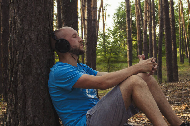 Mężczyzna w słuchawkach siedzi w lesie plecami do drzewa. Pojęcie rekreacji na świeżym powietrzu i relaksu. Dźwięki natury. - Zdjęcie, obraz
