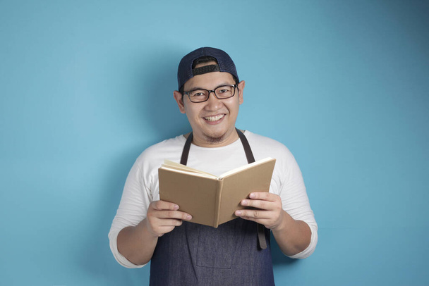 Portrait of Asian male chef reading book of recipes, happy smiling expression, against blue background - Fotografie, Obrázek