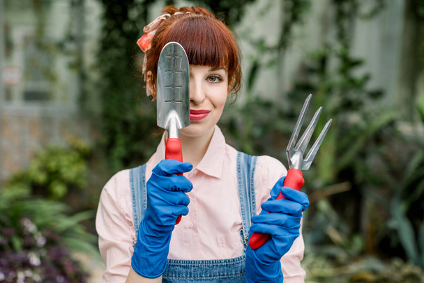 Greenhouse and planting concept. Beautiful young red haired girl, working in hothouse, standing with gardening tools, posing at camera, hiding her eye with spade. - Фото, изображение