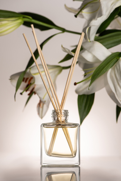 selectieve focus van houten stokken in parfum in fles in de buurt van lelie bloemen op beige achtergrond - Foto, afbeelding