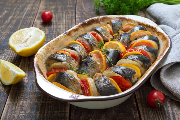 Baked fish with vegetables. Pieces of herring, onions, tomato, lemon, aromatic herbs in ceramic form on a rustic wooden table - Photo, Image