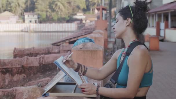 Oldalsó portré gyönyörű női művész dolgozik remekmű szabadban sekély ázsiai háttérrel. Fiatal nő festett vászon képet indiai rakparton lassított felvétel. Boldog szabadidős életmódot! - Felvétel, videó