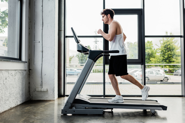 side view of handsome sportsman running on treadmill in gym  - Zdjęcie, obraz