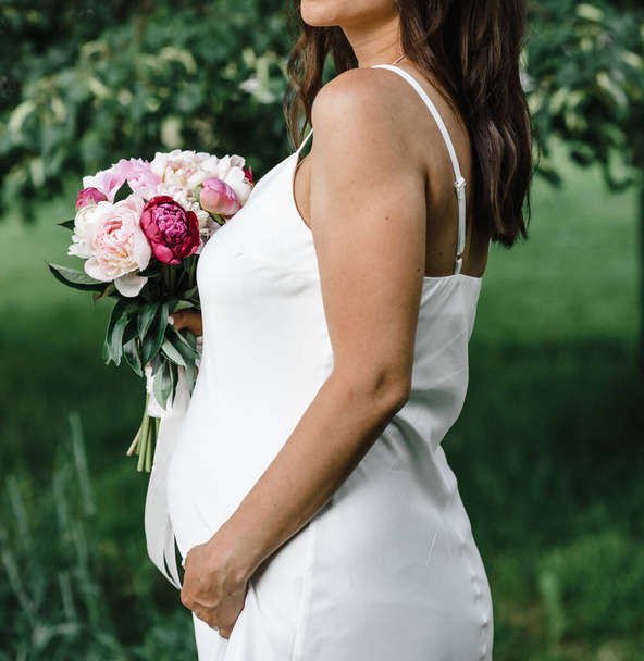 zwangere bruid in een witte dunne jurk op dunne bandjes met blote schouders met een boeket pioenrozen. moederschap - Foto, afbeelding
