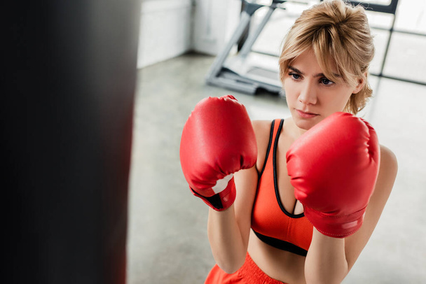 selective focus of young sportswoman in boxing gloves looking at punching bag  - Foto, imagen