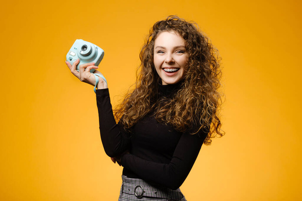 Kıvırcık saçlı, güzel gülüşlü, sarı arka planda izole fotoğraf makinesi olan mutlu bir kız. - Fotoğraf, Görsel