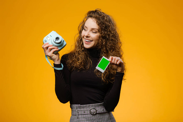 Beautiful girl with instant film camera isolated on yellow background. Model is holding blank frame with mockup space on a picture - Photo, Image