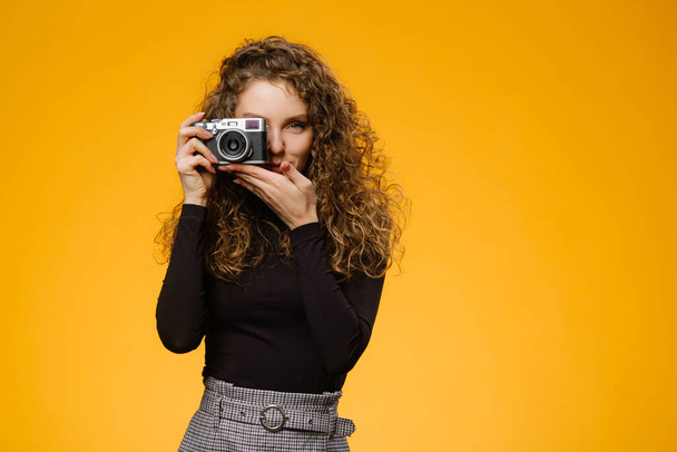 Beautiful girl with photo camera looking at viewfinder and taking a shot. isolated on yellow background - Fotoğraf, Görsel