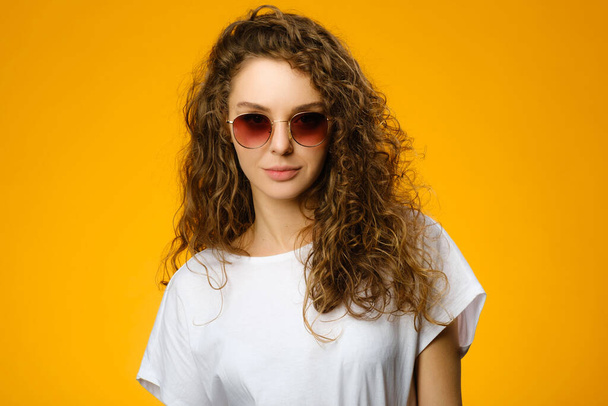 Pretty teenager with sunglasses and white shirt looking at camera. Model isolated on colorful yellow background - Photo, Image
