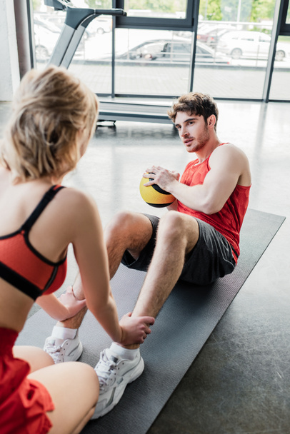 selective focus of sportive woman holding legs of sportsman exercising with ball on fitness mat  - Photo, Image