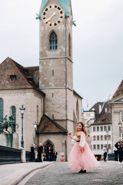 a little girl in a pink Princess dress with a bouquet in her hands walks through the old city of Zurich.Portrait of a girl in a pink dress on a city street in Switzerland. - 写真・画像