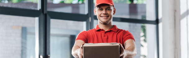 panoramiczne ujęcie szczęśliwego dostawcy trzymającego pudełko kartonowe  - Zdjęcie, obraz