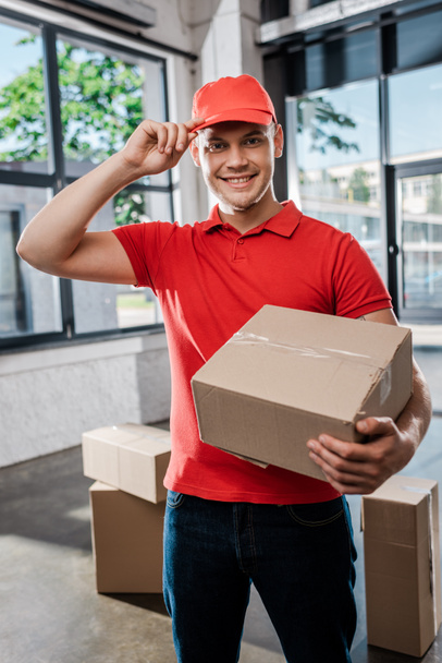 happy delivery man touching cap while holding carton box - Φωτογραφία, εικόνα