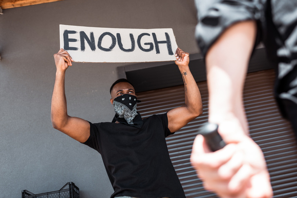 foyer sélectif de l'homme afro-américain avec écharpe sur le visage tenant la plaque avec assez de lettrage près de policier avec matraque, concept de racisme  - Photo, image