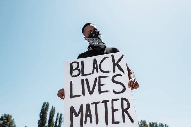 low angle view of african american man with scarf on face holding placard with black lives matter lettering against sky - Foto, imagen