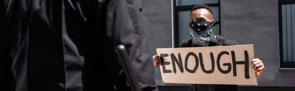 selective focus of african american man with scarf on face holding placard with enough lettering near policeman with baton on street, racism concept  - Photo, Image