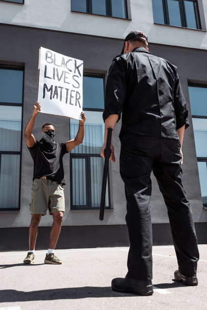 selective focus of african american man with scarf on face holding placard with black lives matter lettering near policeman with baton on street, racism concept  - Foto, immagini