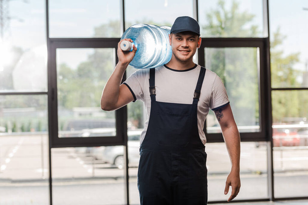 cheerful delivery man in cap and uniform holding gallon of water in bottle  - Photo, Image