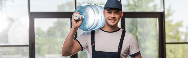 panoramic concept of cheerful delivery man in cap and uniform holding bottled water  - Φωτογραφία, εικόνα