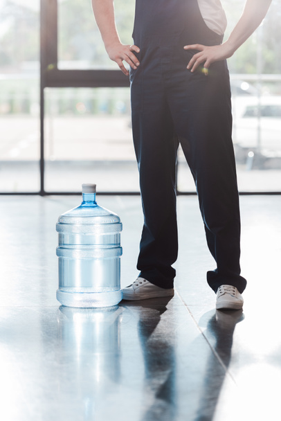 Ausgeschnittene Ansicht eines Lieferanten in Uniform, der mit Händen auf Hüften in der Nähe von abgefülltem Wasser steht  - Foto, Bild