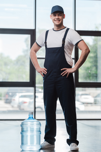 fröhlicher Auslieferer in Uniform, der mit den Händen auf den Hüften neben abgefülltem Wasser steht  - Foto, Bild