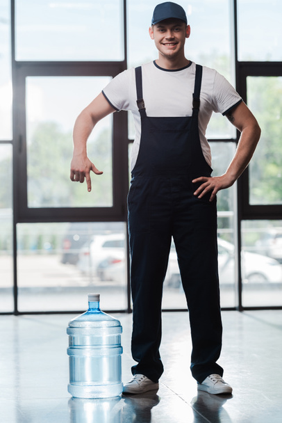 веселий чоловік доставки в уніформі, що стоїть з рукою на стегні і вказує пальцем у пляшковій воді
  - Фото, зображення