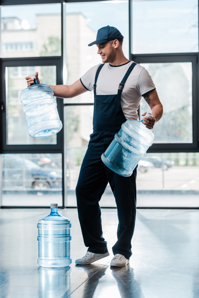 vrolijke bezorger in uniform kijkend naar lege flessen in de buurt van gallon water  - Foto, afbeelding