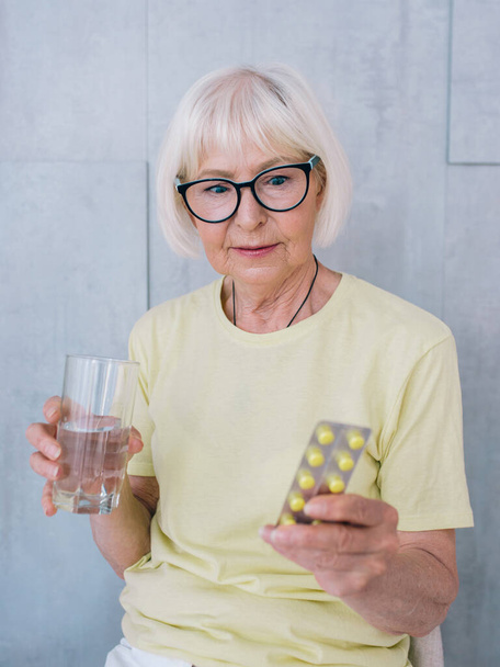 senior woman in glasses holding medicine (pills) and glass of water. Age, health care, treatment concept - Photo, Image