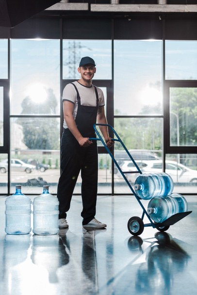 cheerful delivery man in uniform holding hand truck with bottled water  - Fotoğraf, Görsel