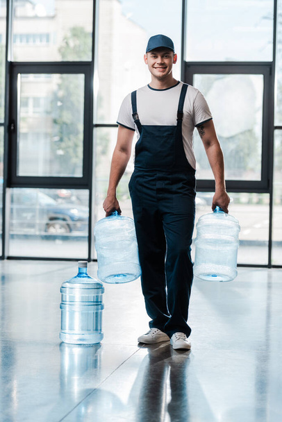 cheerful delivery man in uniform holding empty bottles near gallon of water  - Fotoğraf, Görsel