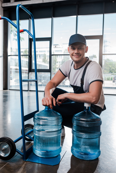 happy delivery man in uniform sitting near hand truck with purified water in bottles  - Valokuva, kuva
