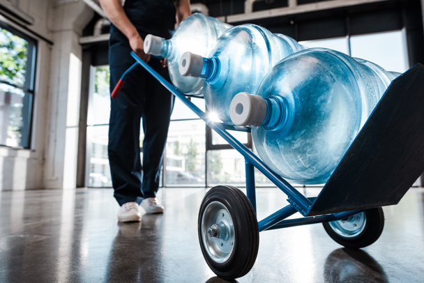 cropped view of delivery man in uniform holding hand truck with purified water in bottles - Fotografie, Obrázek