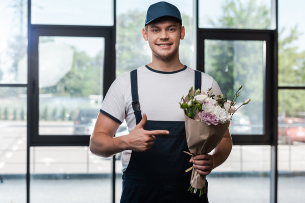 happy delivery man in uniform en pet wijzend met de vinger naar boeket met bloemen  - Foto, afbeelding