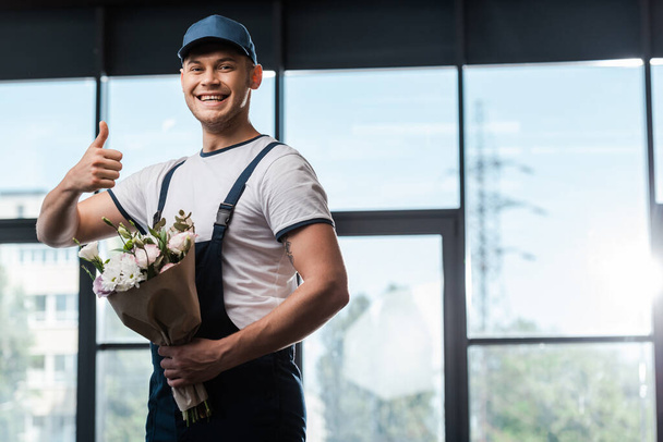 homme de livraison heureux en uniforme et chapeau montrant pouce vers le haut et tenant bouquet avec des fleurs  - Photo, image