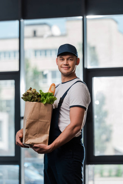 cheerful delivery man in cap holding paper bag with groceries  - 写真・画像