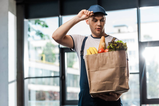 surprised delivery man touching cap and looking at paper bag with groceries  - Fotoğraf, Görsel