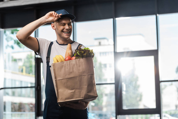 happy delivery man touching cap and holding paper bag with groceries  - Photo, Image