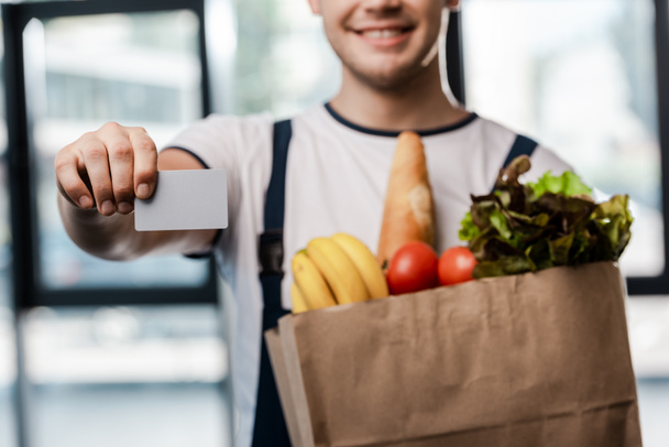 cropped view of happy delivery man holding paper bag with groceries and blank card - Foto, afbeelding