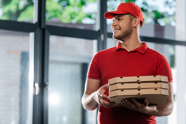 happy delivery man in cap looking away and holding pizza boxes  - Φωτογραφία, εικόνα