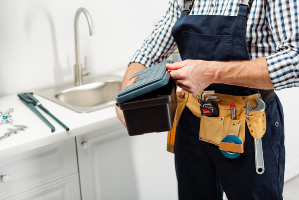 Cropped view of plumber with tools on tool belt holding toolbox in kitchen  - Fotó, kép